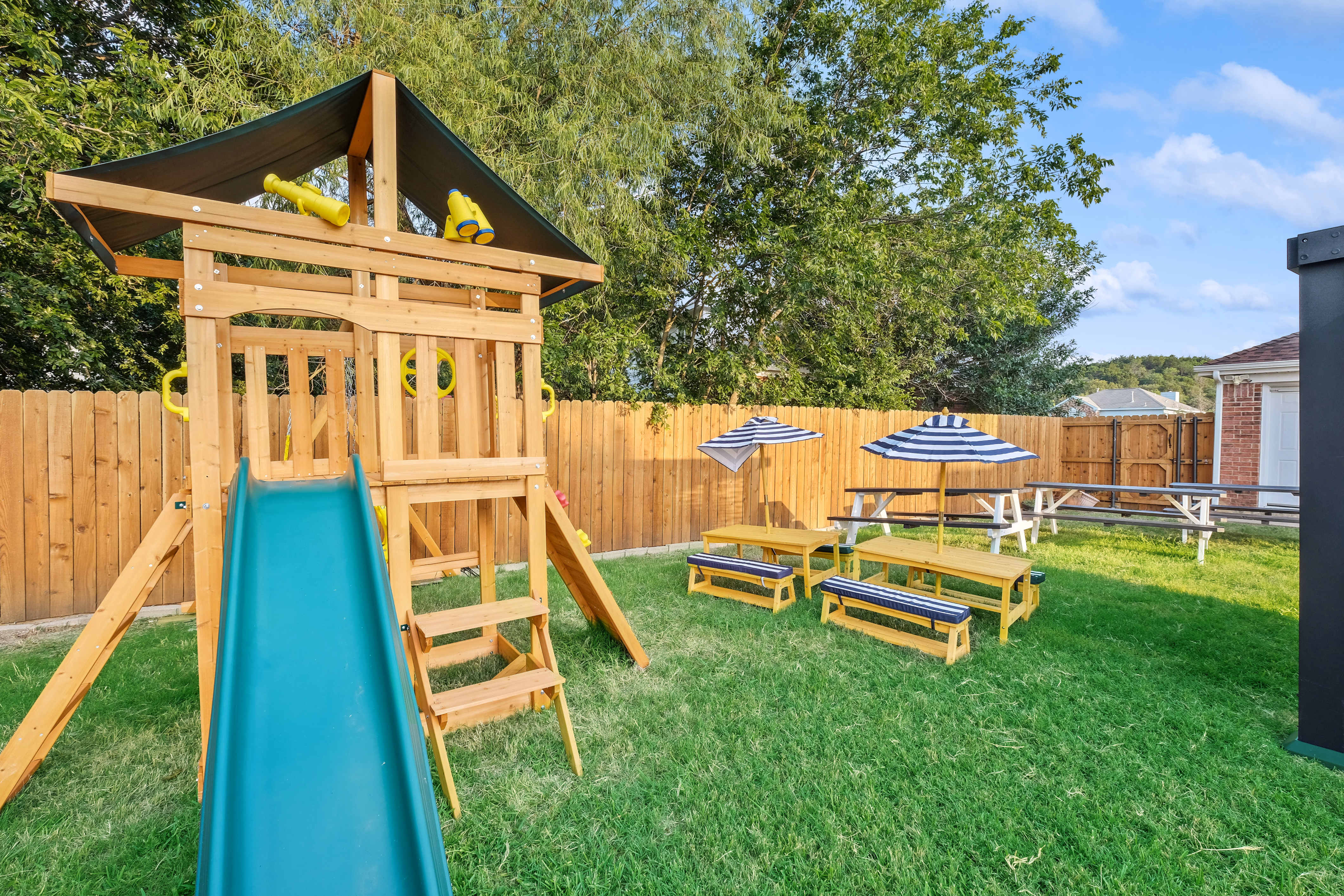 Wooden playset with green slide and striped picnic tables.